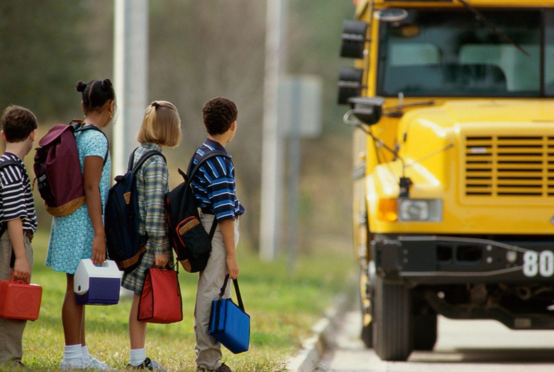 Four children with backpacks and lunch boxes waiting for a school bus.