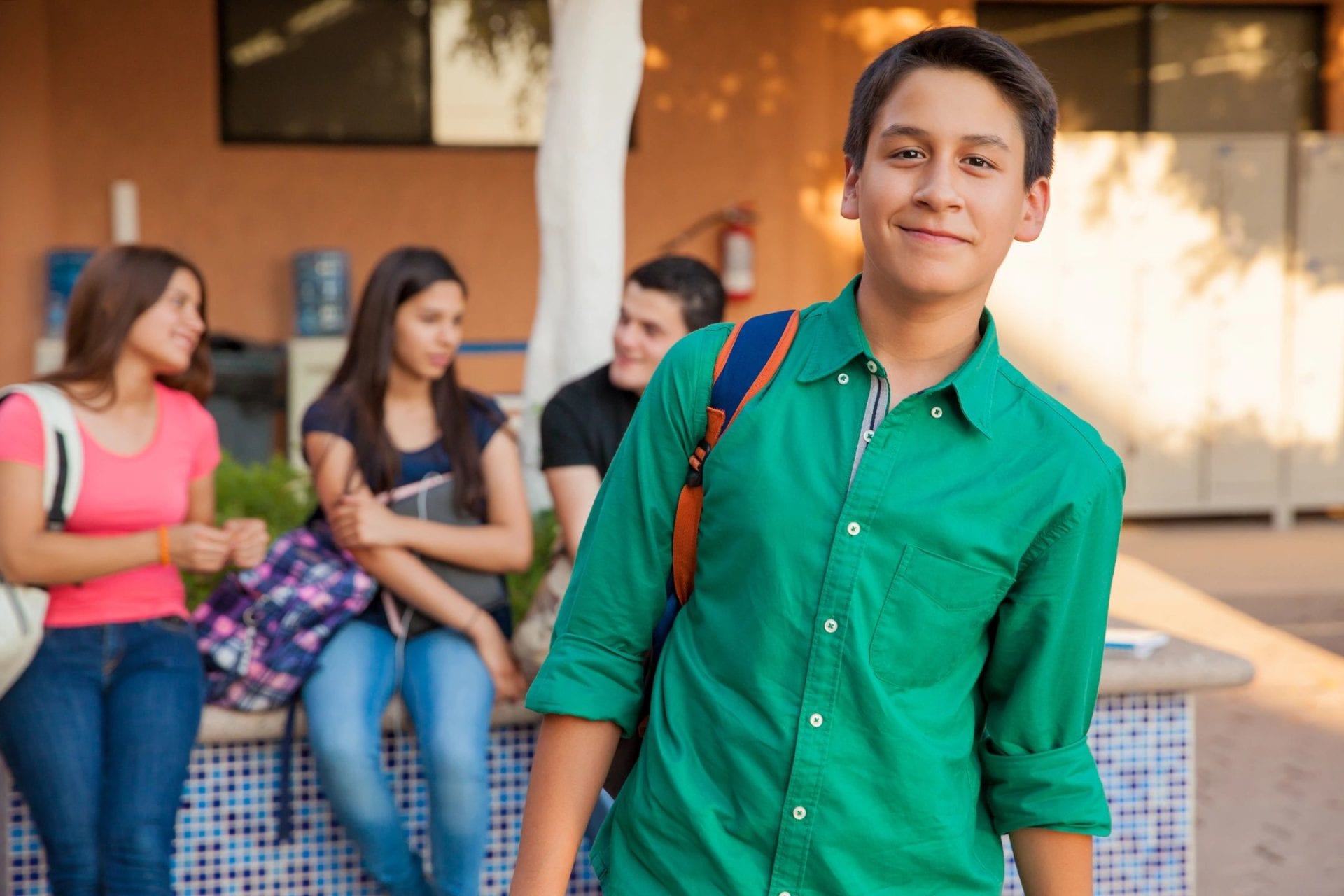 A smiling teenage boy with a backpack outside a school, with classmates talking behind him.