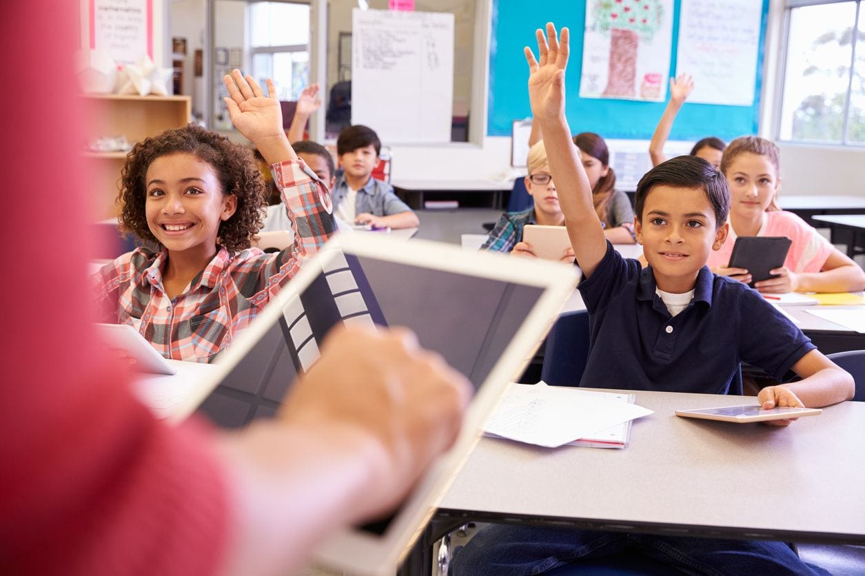 Elementary students raising their hands in a classroom.