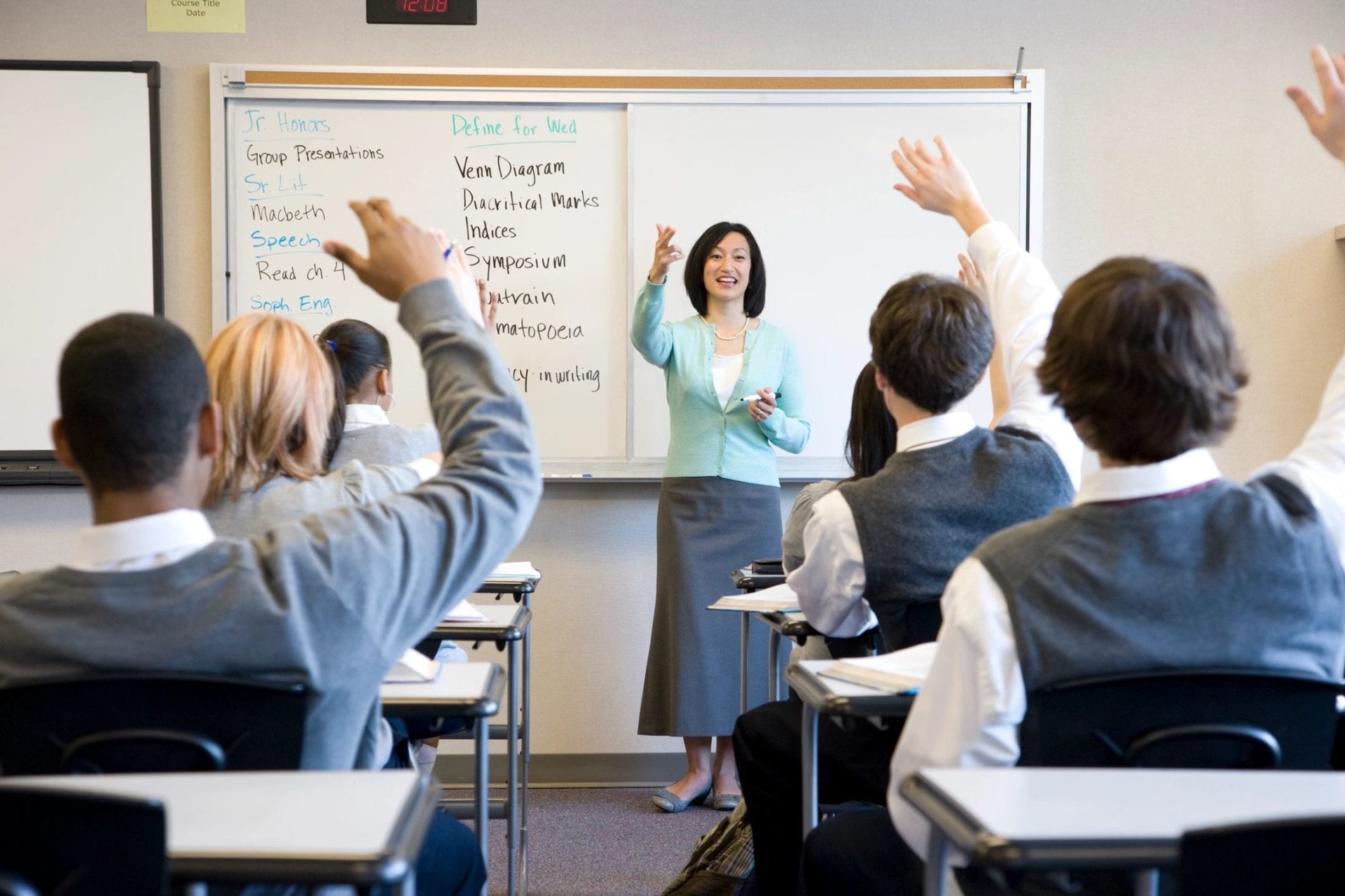 A teacher at a whiteboard calls on students with raised hands.