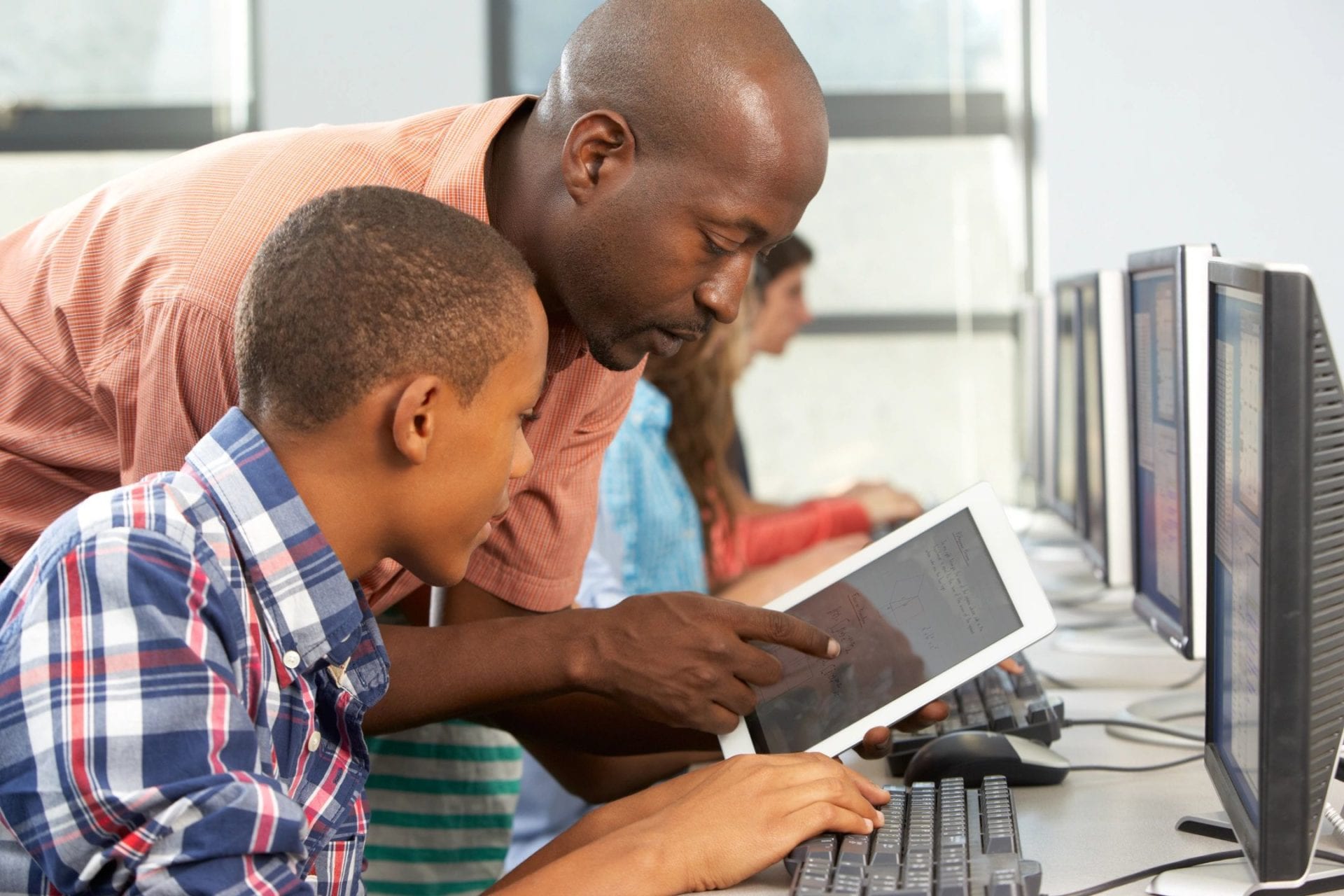 A teacher points to a tablet while a student works at a computer.